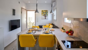 A modern kitchen with a dining table set for two, a TV mounted on the wall, and a fruit basket on the counter. - White Rock Milos Suites (Milos)