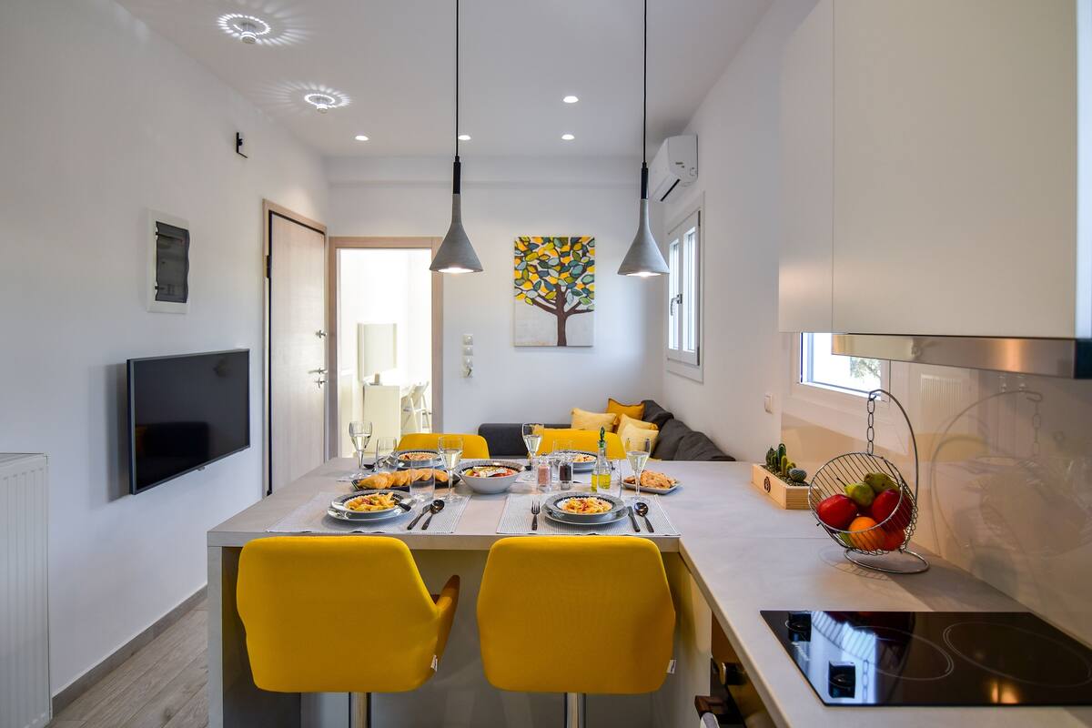 A modern kitchen with a dining table set for two, a TV mounted on the wall, and a fruit basket on the counter.