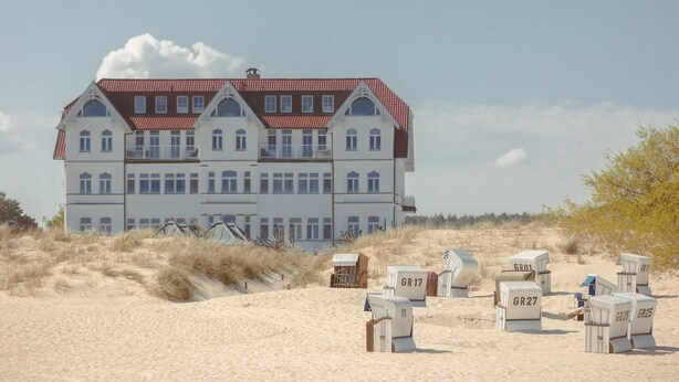 A large, multi-story building with a red-tiled roof and white walls, situated on a sandy beach with dunes.