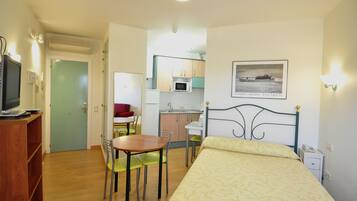 Una habitación pequeña con cama, escritorio, silla, televisión y kitchenette.