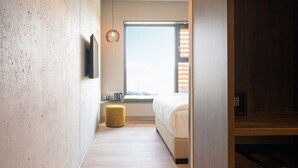 Habitación de hotel moderna con suelo de madera, una ventana grande, una cama, una mesita pequeña y un televisor montado en la pared.