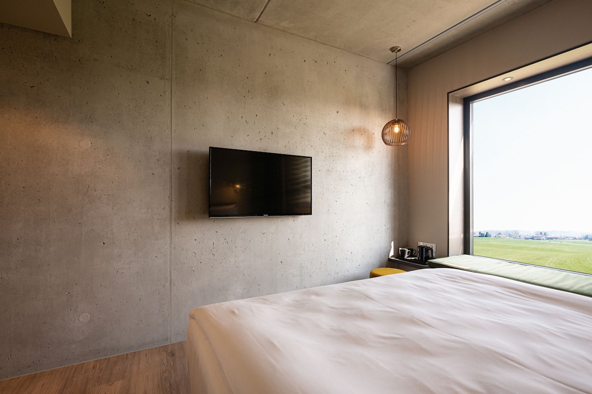 Habitación de hotel moderna con una cama grande, un televisor de pantalla plana en la pared, una mesita con una taza y una tetera, y un ventanal con vistas a un campo verde.