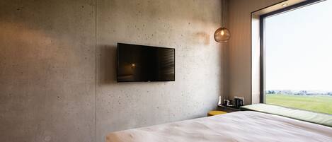 Habitación de hotel moderna con una cama grande, un televisor de pantalla plana en la pared, una mesita con una taza y una tetera, y un ventanal con vistas a un campo verde.