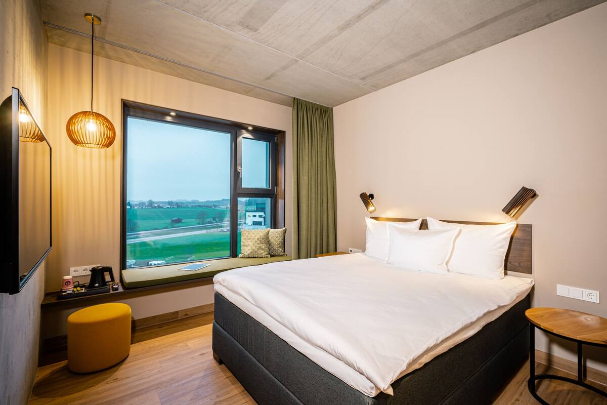 Una habitación de hotel moderna con una cama grande, un ventanal con vistas a un paisaje verde, una mesita pequeña y un banco.
