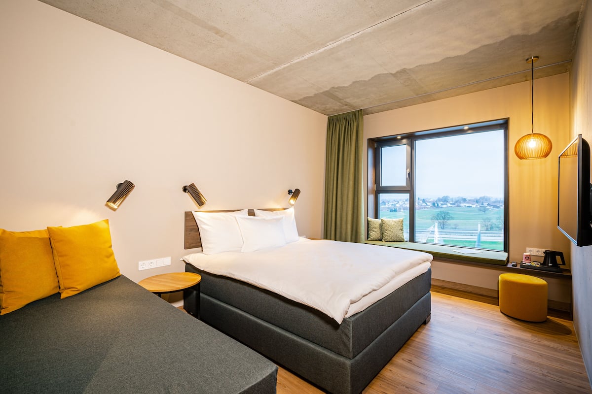 Habitación de hotel moderna con una cama grande, un sofá, una mesita y una ventana con vistas.