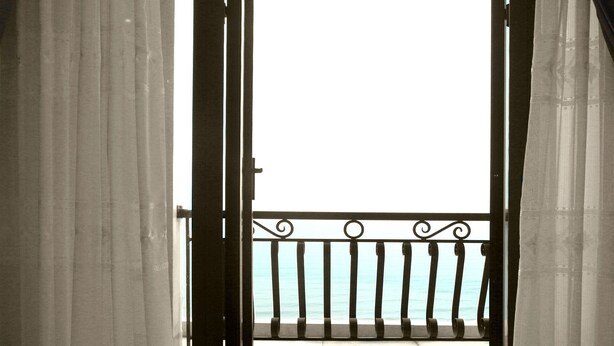 Een open deur die leidt naar een balkon met een metalen hek en uitzicht op de zee.