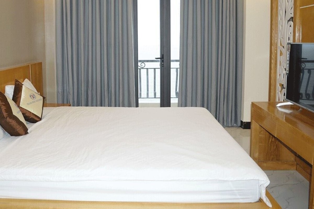 Ein Hotelzimmer mit einem großen Bett, einem Holz-Kopfbrett, einem Flachbildfernseher auf einem Holzboden und einem Balkon mit grauen Vorhängen.