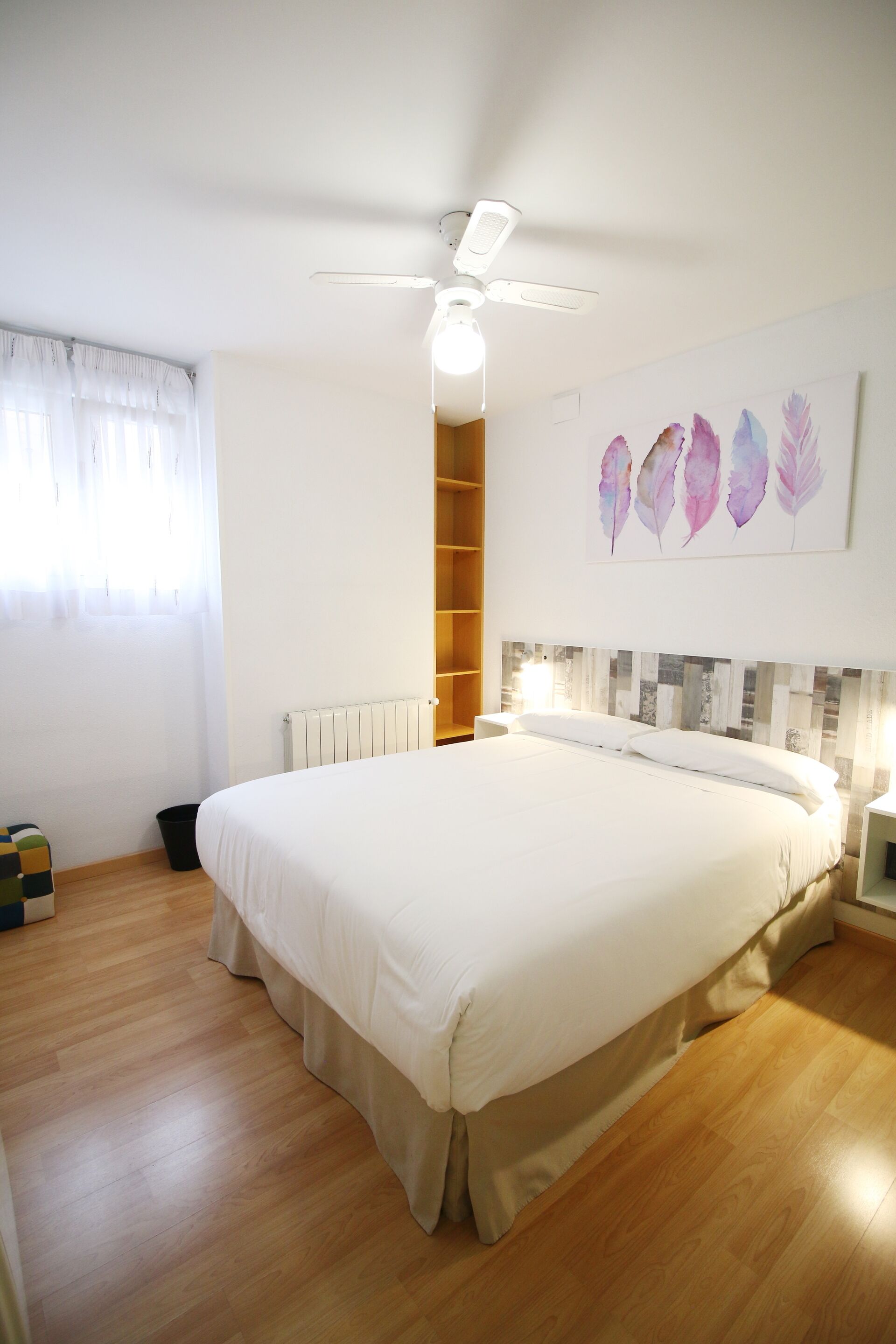 A bedroom with a large bed, a ceiling fan, a radiator, and a wall with a feather artwork.