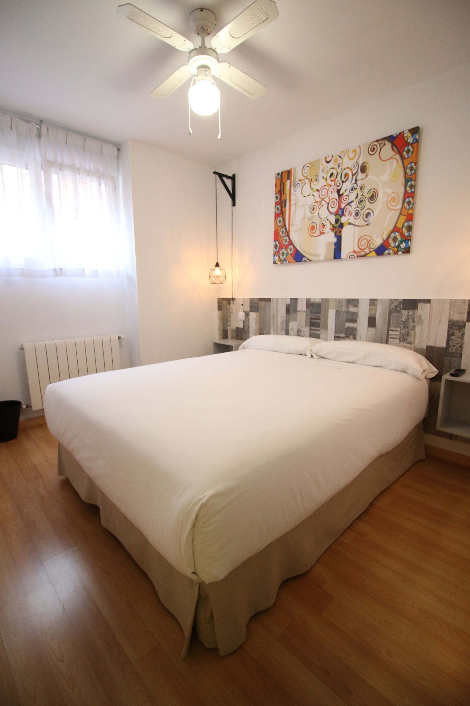 A bedroom with a large bed, a ceiling fan, a radiator, a wall-mounted light, and a colorful abstract painting.