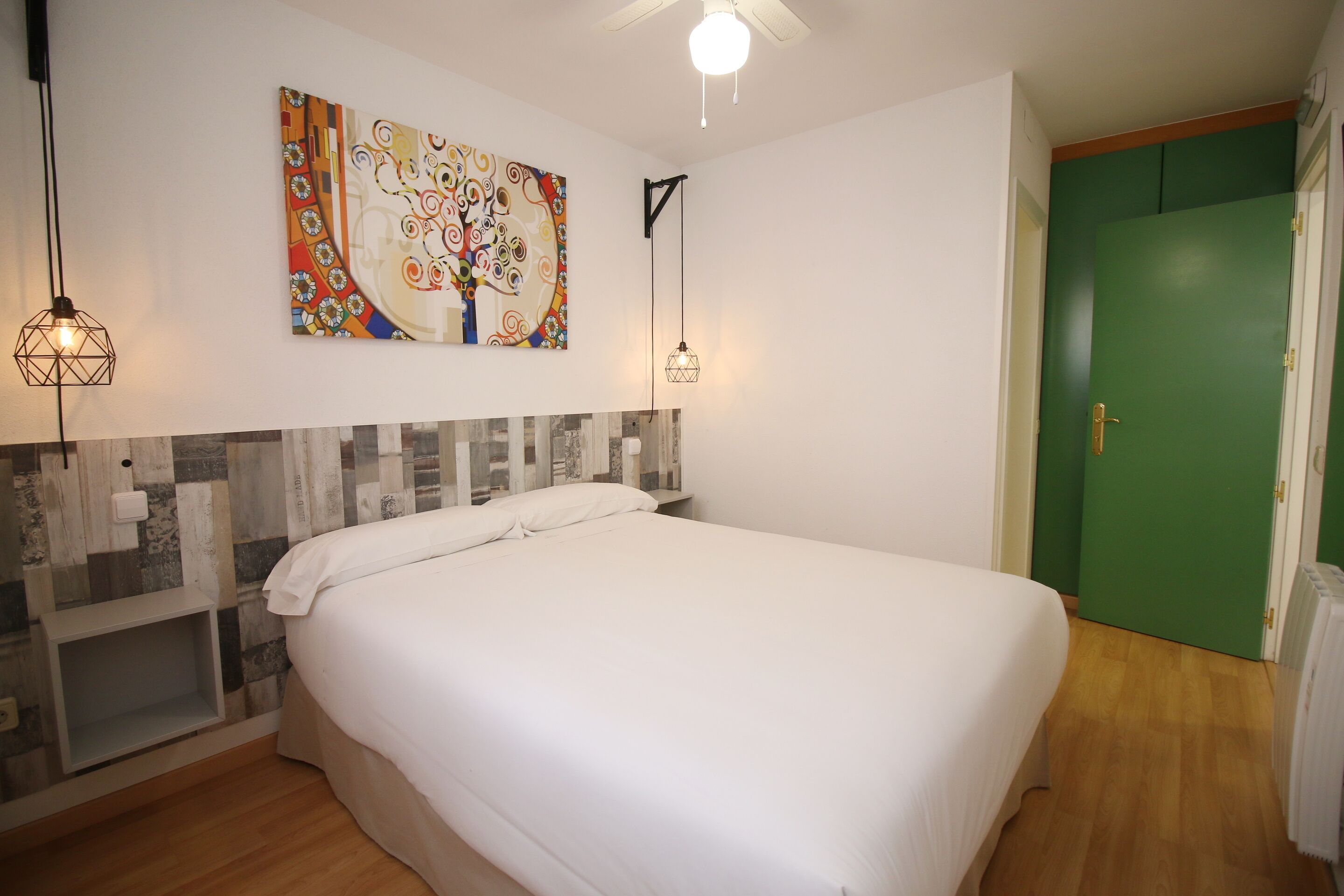 Un dormitorio con una cama grande, un cuadro abstracto de colores, dos lámparas colgantes y una puerta verde.