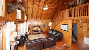 L’intérieur d’un chalet en bois, avec une tête de wapiti accrochée au mur, un ventilateur de plafond, un téléviseur, un canapé et un escalier.