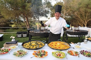 A chef standing behind a table with large pans of food, surrounded by various dishes and outdoor dining furniture.