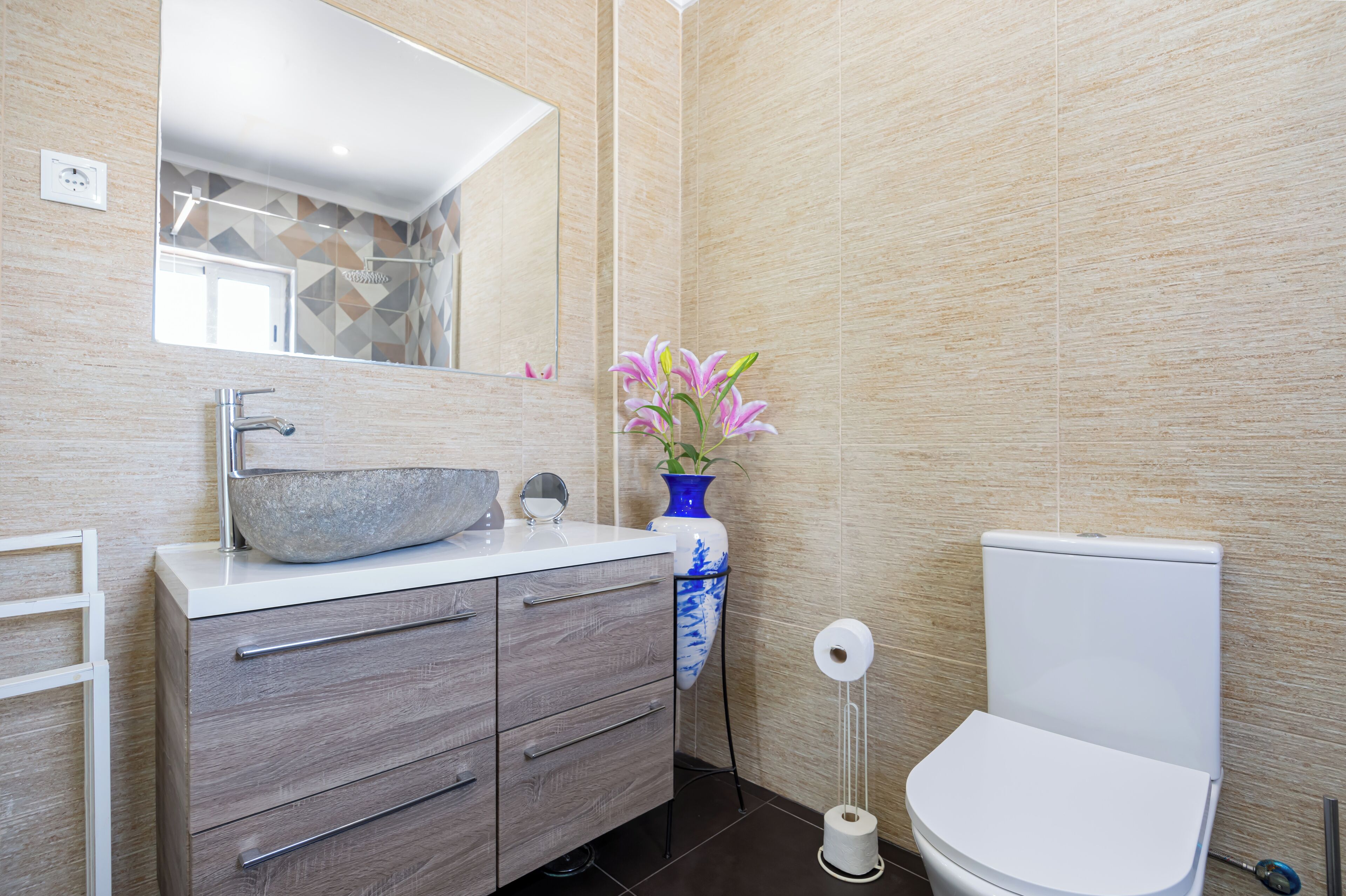 A modern bathroom with a large mirror, a stone sink, a blue vase with flowers, and a toilet.