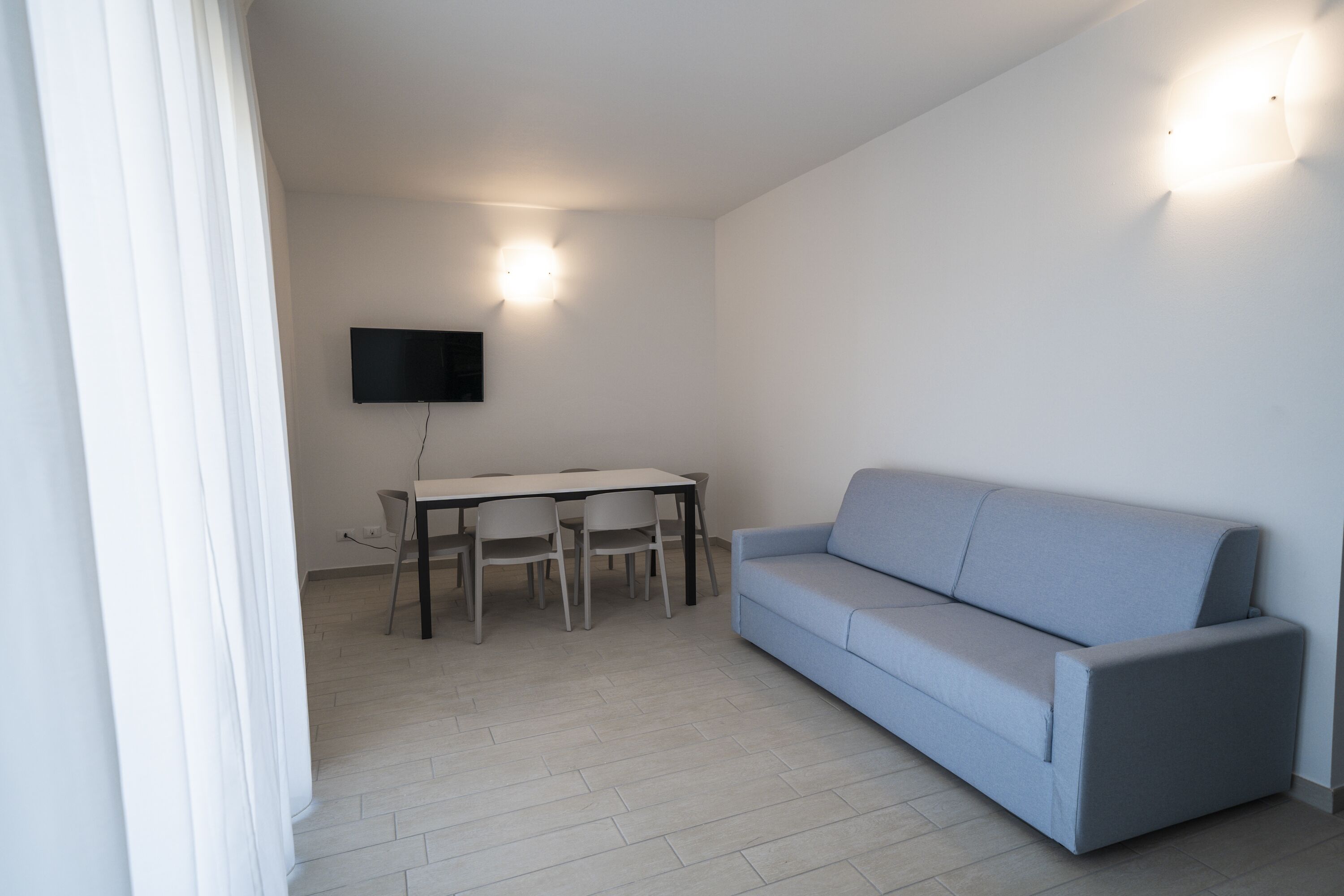 A modern living room with a light blue sofa, a dining table with chairs, and a flat-screen TV mounted on the wall.