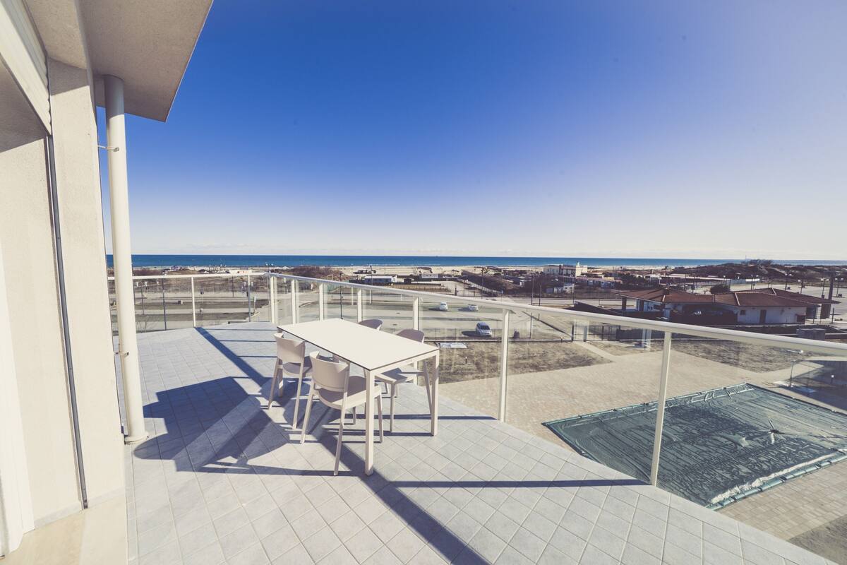 Un balcón con mesa y sillas, con vistas a una playa y un estacionamiento.