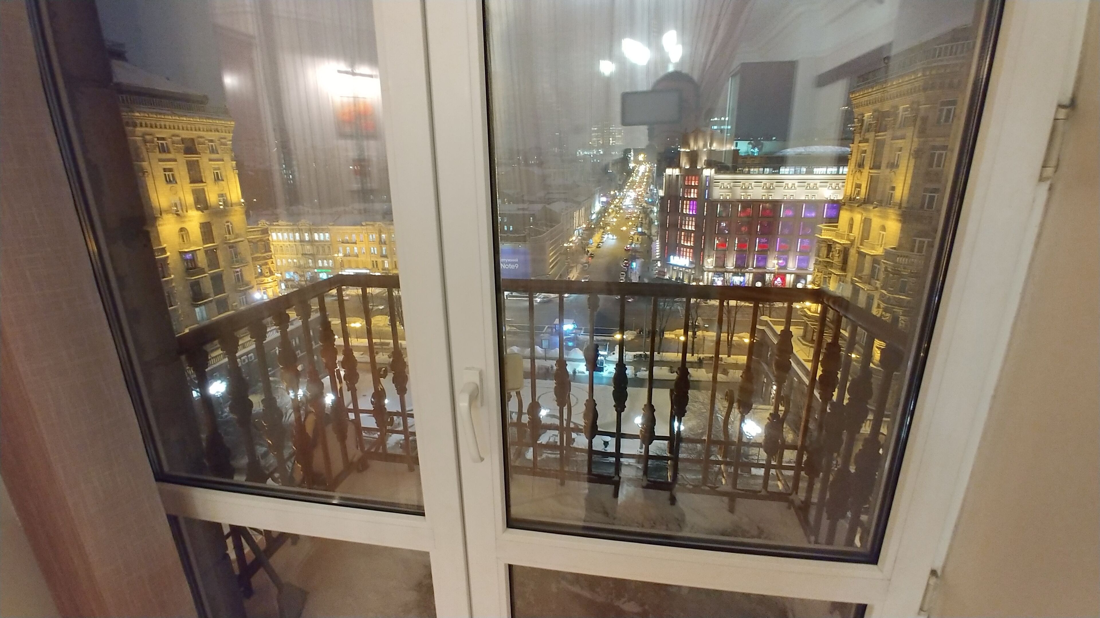Näkymä ikkunasta, jossa on metallinen kaide. Ikkunasta avautuu näkymä kaupungin kadulle yöllä, ja näkyvissä on valaistuja rakennuksia ja vilkasliikenteinen tie.