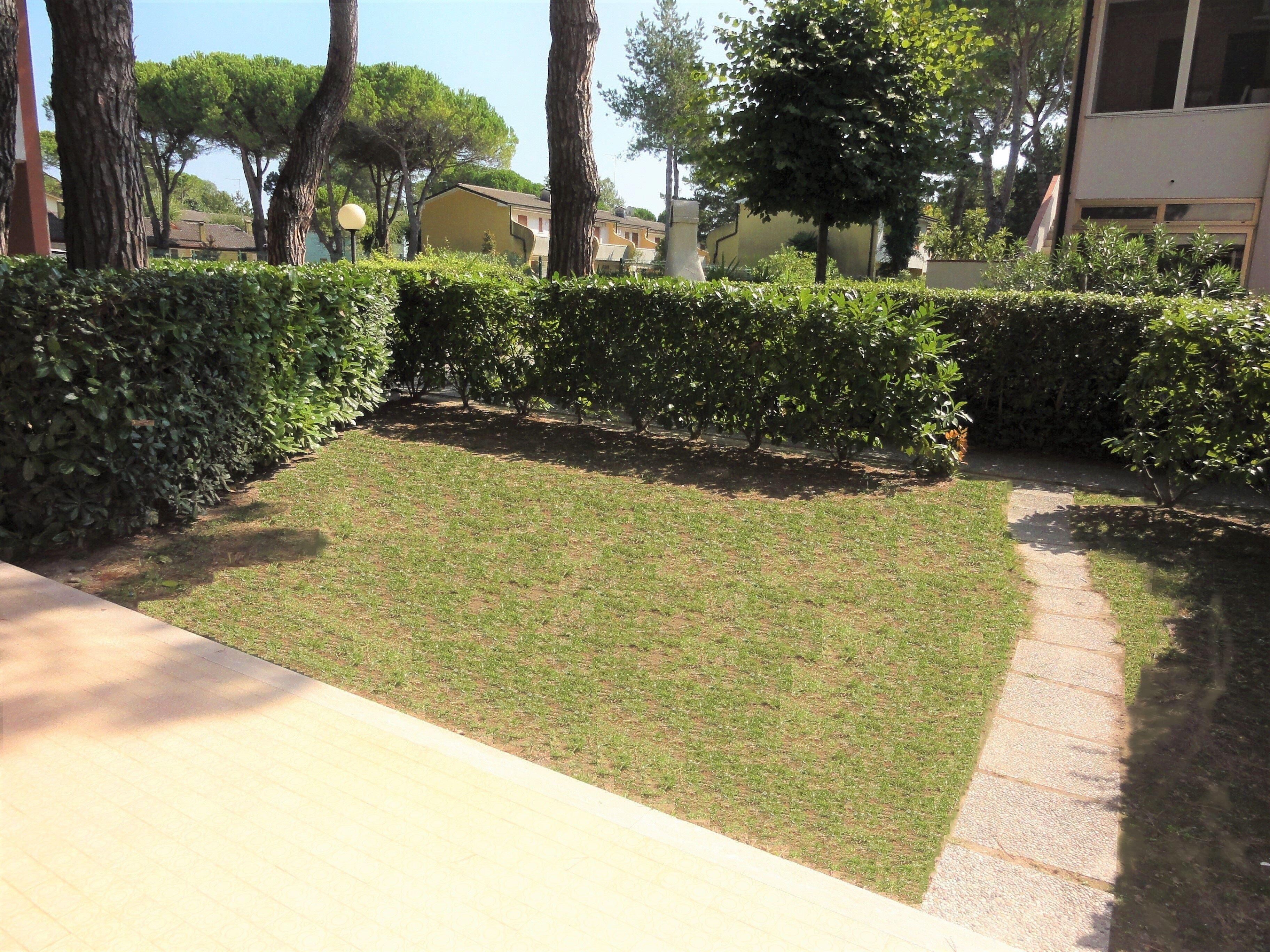 A well-maintained grassy area with a paved walkway, surrounded by neatly trimmed hedges and trees.