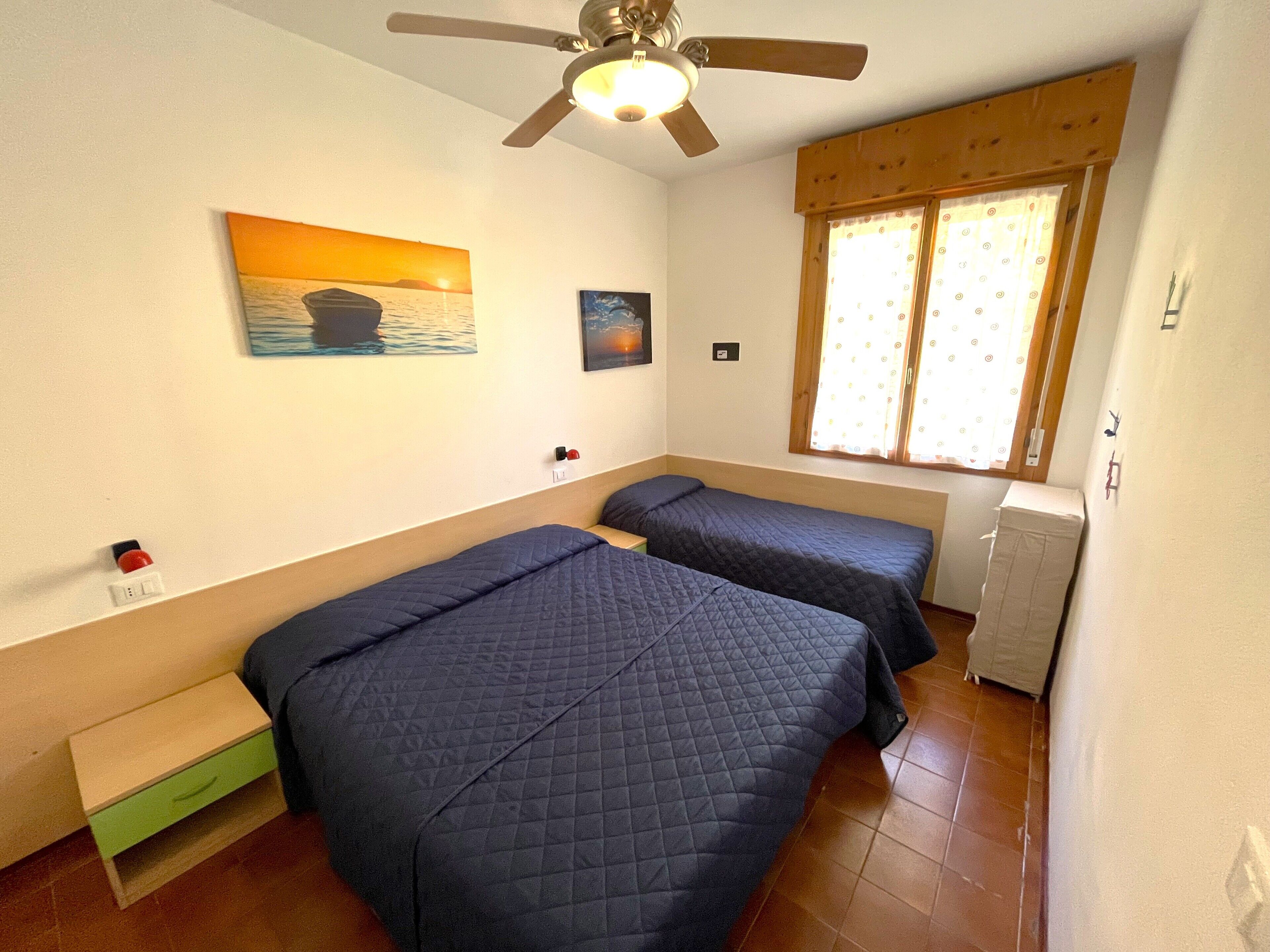 A bedroom with two beds, a ceiling fan, a window with curtains, and a painting on the wall.