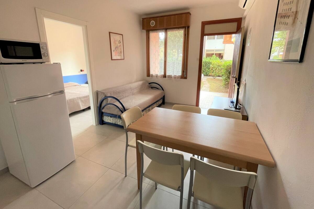 Une pièce compacte comprenant un coin cuisine, un lit simple, une petite table avec deux chaises et une fenêtre avec des rideaux.