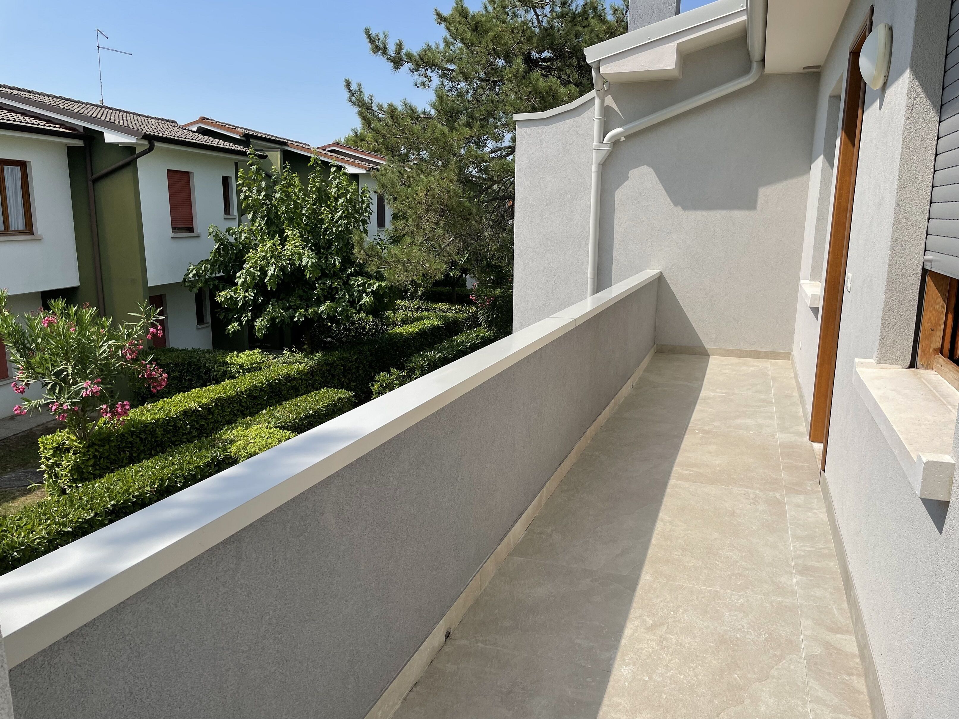Ein Wohnbalkon mit Fliesenboden, weißer Wand und einer Hecke mit rosa Blüten.