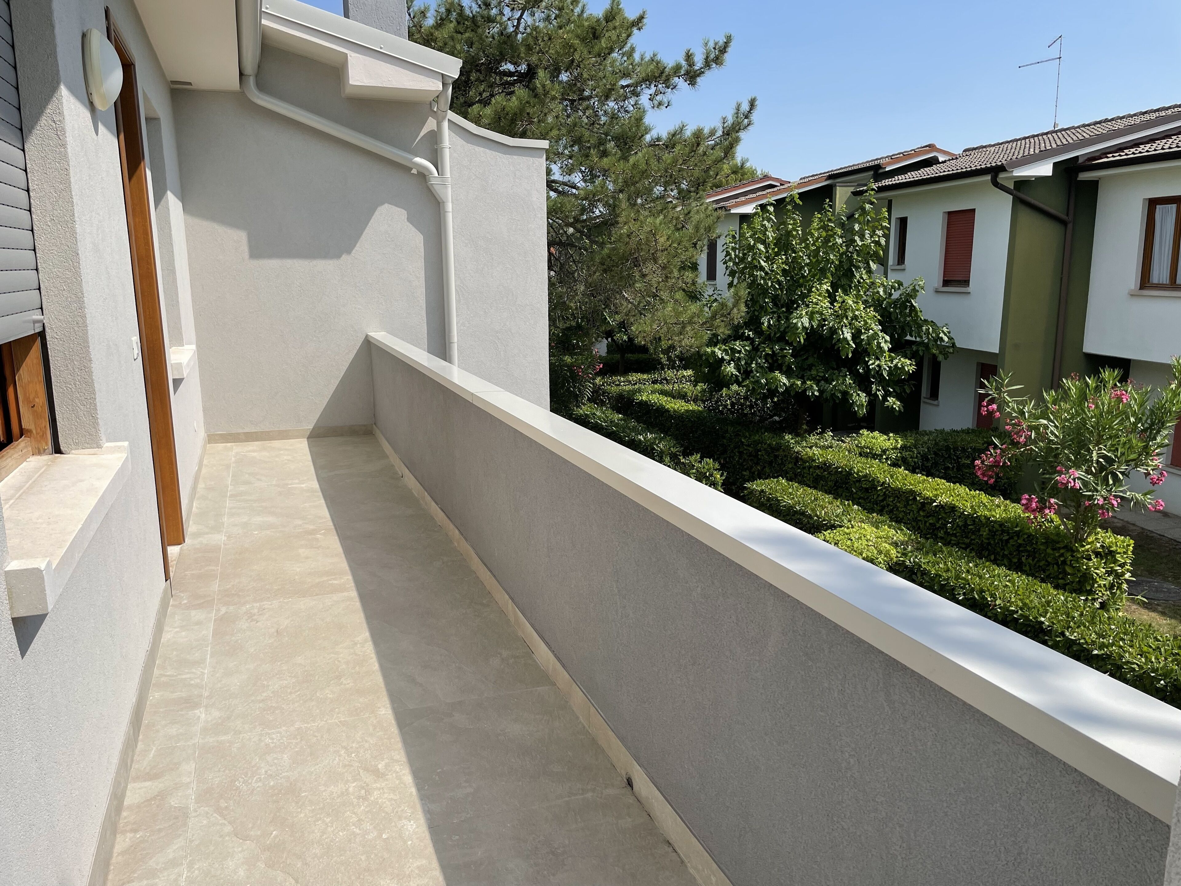 Un balcon étroit, doté d’un sol carrelé, jouxtant un bâtiment à la façade blanche et muni d’une fenêtre à cadre en bois.