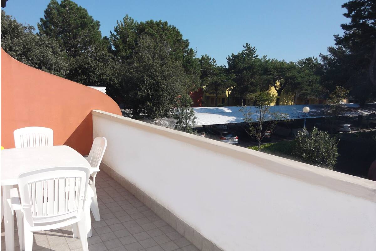 Un balcon avec une table et des chaises blanches, un sol carrelé et une vue sur les arbres et les voitures garées.