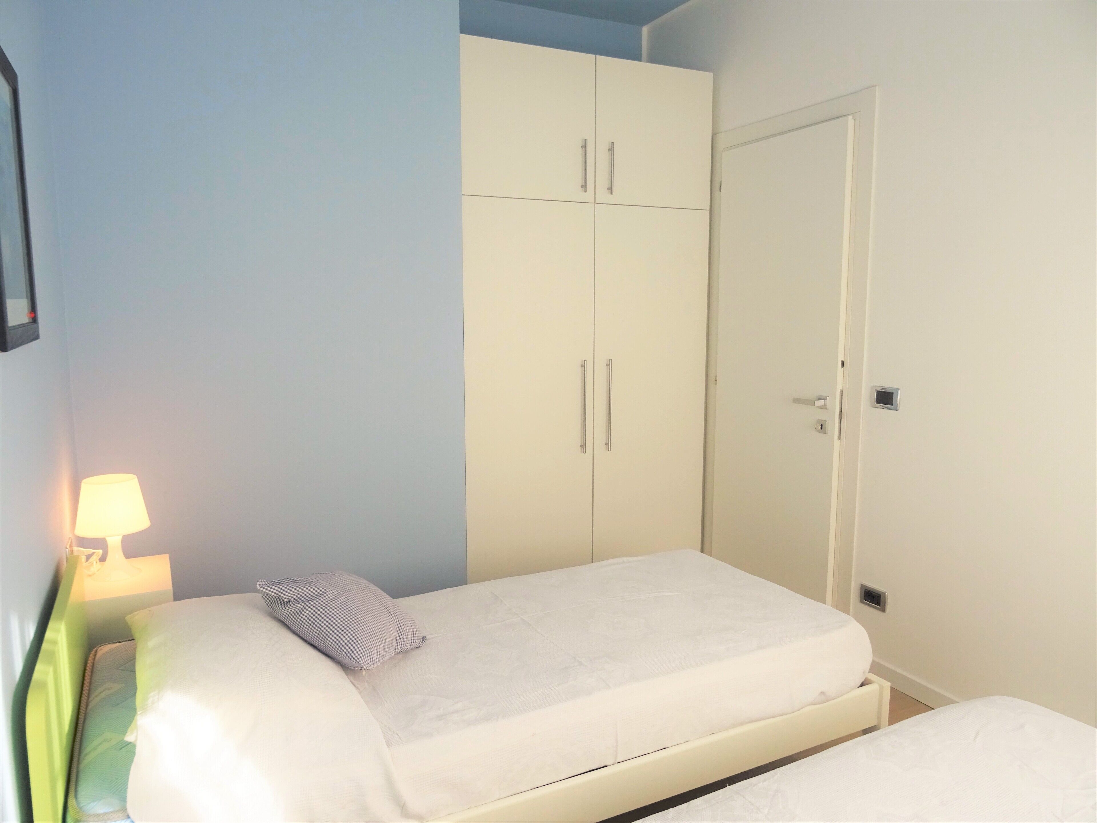 A bedroom with a bed, a nightstand, a lamp, a wardrobe, and a door.