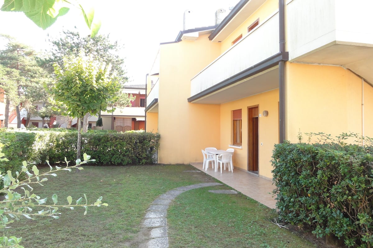 Une cour avec un petit jardin, une table et des chaises blanches, et un bâtiment jaune doté d’un balcon.