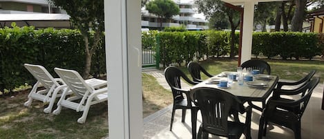 Um pátio externo com mesa e cadeiras cobertas, rodeado por vegetação e com vista para edifícios ao fundo.