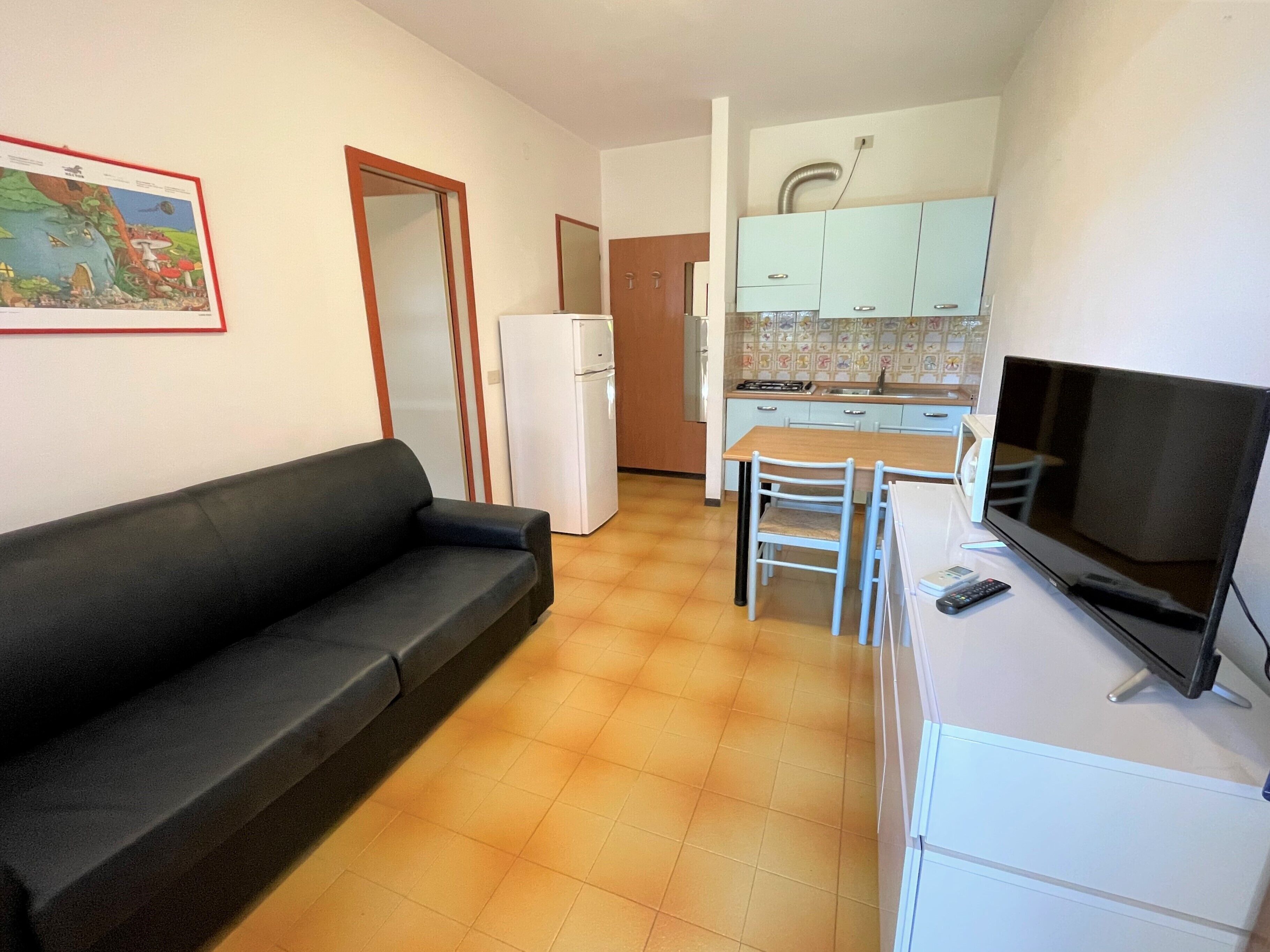 Ein gemütlicher Wohnraum mit Sofa, Fernseher, Esstisch mit Stühlen, Küchenzeile inklusive Schränke und Kühlschrank.