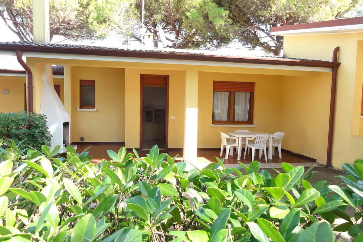 Un bâtiment jaune avec un patio couvert, une porte et une fenêtre avec des rideaux. On y trouve des chaises et une table en plastique, toutes deux blanches, sur le patio.