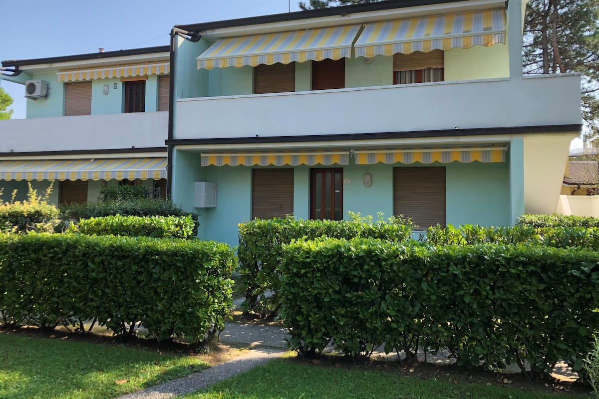 Un bâtiment à deux étages, doté d’une façade vert clair, d’auvents à rayures jaunes et blanches et d’un petit jardin où les arbustes sont soigneusement taillés.
