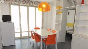 Une pièce avec une table blanche et des chaises orange, un meuble blanc et un téléviseur posé sur un autre meuble blanc.
