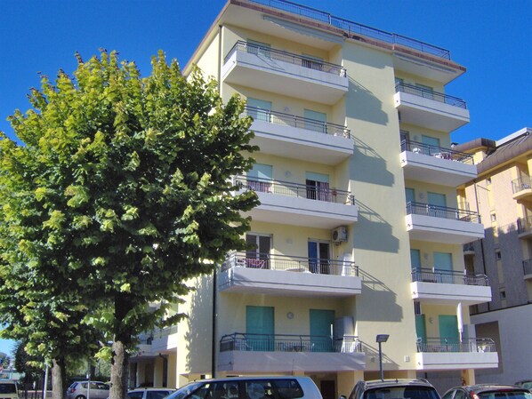 A multi-story residential building with balconies, parked cars, and a large tree in front.