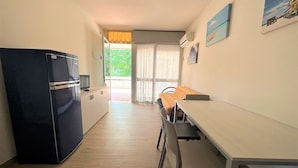 Une cuisine compacte équipée d’un réfrigérateur, d’une table et de chaises. Une porte-fenêtre coulissante donne accès à un espace extérieur.