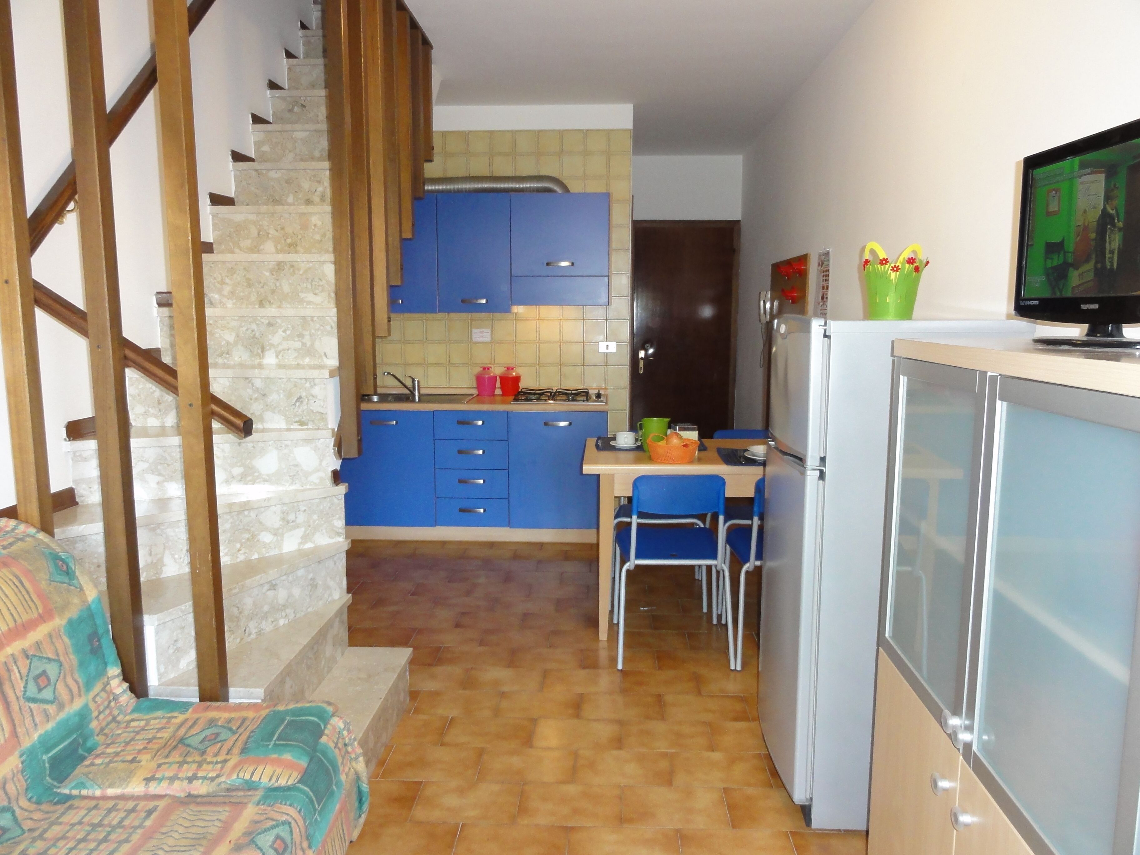 A compact kitchen with blue cabinets, a small dining area with two chairs, and a staircase leading to an upper level.