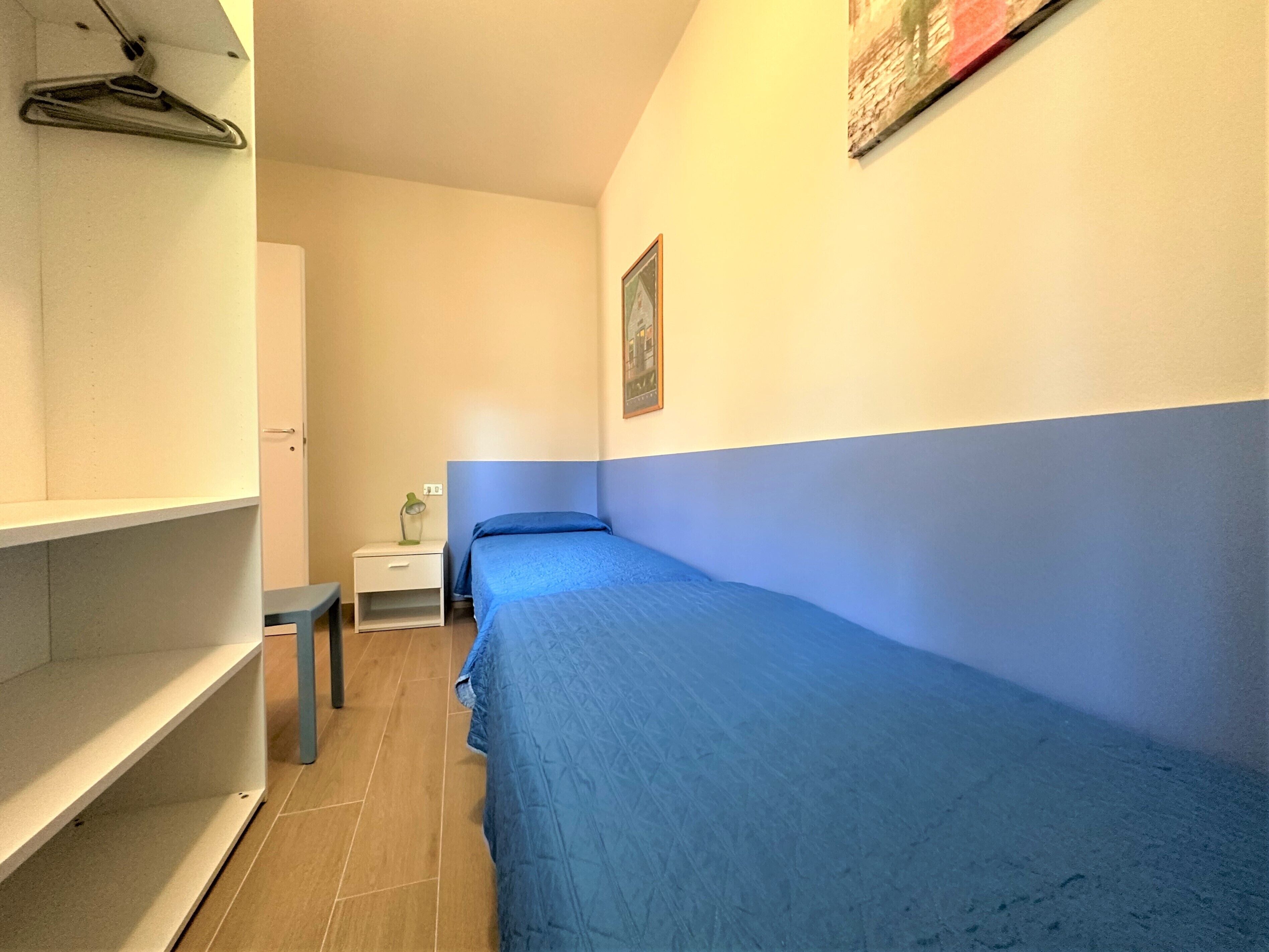 A narrow room with a single bed, a nightstand, a small table, and a hanging coat rack.