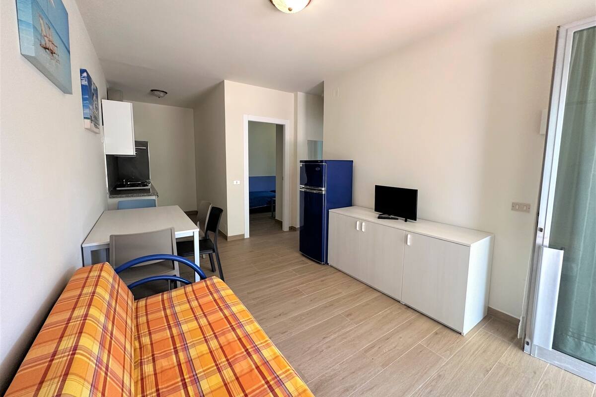 Ein gemütlicher Wohnbereich mit Küchenzeile, Essplatz, einem Bett mit Karoüberwurf und einem Fernseher auf einem Schrank.