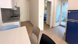 Une cuisine compacte équipée d’une cuisinière, d’une table à manger avec des chaises et d’un réfrigérateur. Un couloir mène à une chambre avec un lit et à une salle de bain avec des toilettes.