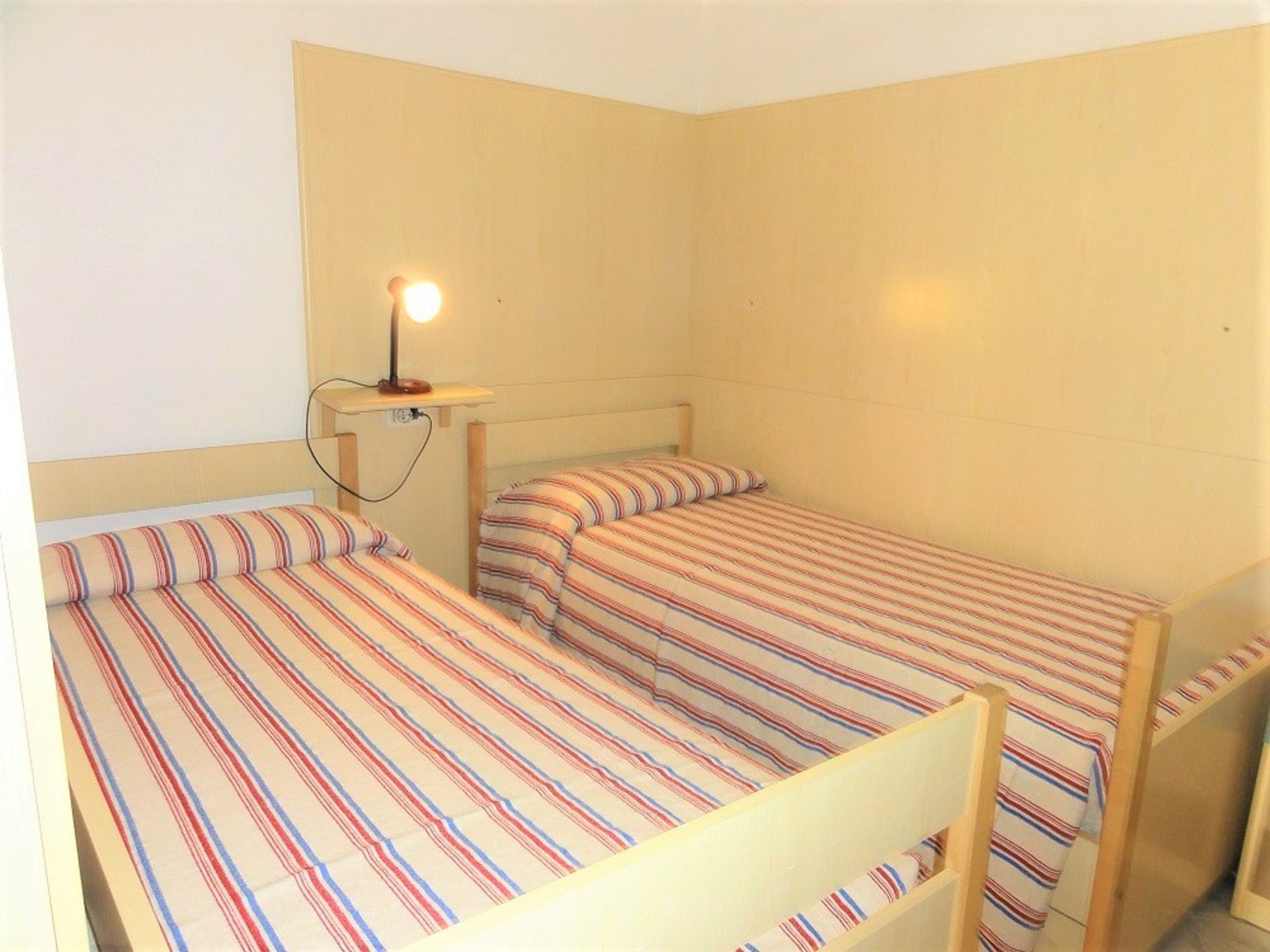 Un petit logement composé d’une seule pièce, avec deux lits, une table de chevet avec une lampe et des murs aux couleurs neutres.