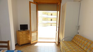 A room with a bed, a TV on a wooden dresser, a table with a blue tablecloth, and a view of a building through an open door. - Condominio Ausonia Al Mare (San Michele al Tagliamento)