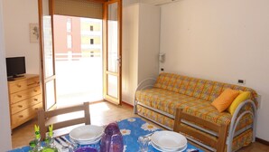 A small, bright room with a wooden table set for dining, a sofa, and a TV on a wooden cabinet. - Condominio Ausonia Al Mare (San Michele al Tagliamento)
