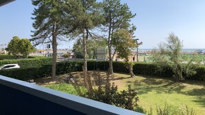 Une vue depuis une fenêtre donnant sur un espace verdoyant avec des arbres, un panneau de signalisation et, au loin, une plage parsemée de parasols.