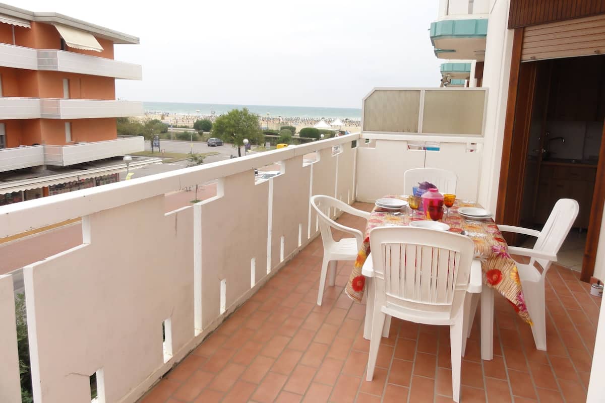 A balcony with a table set for two, white plastic chairs, and a view of the beach and sea.