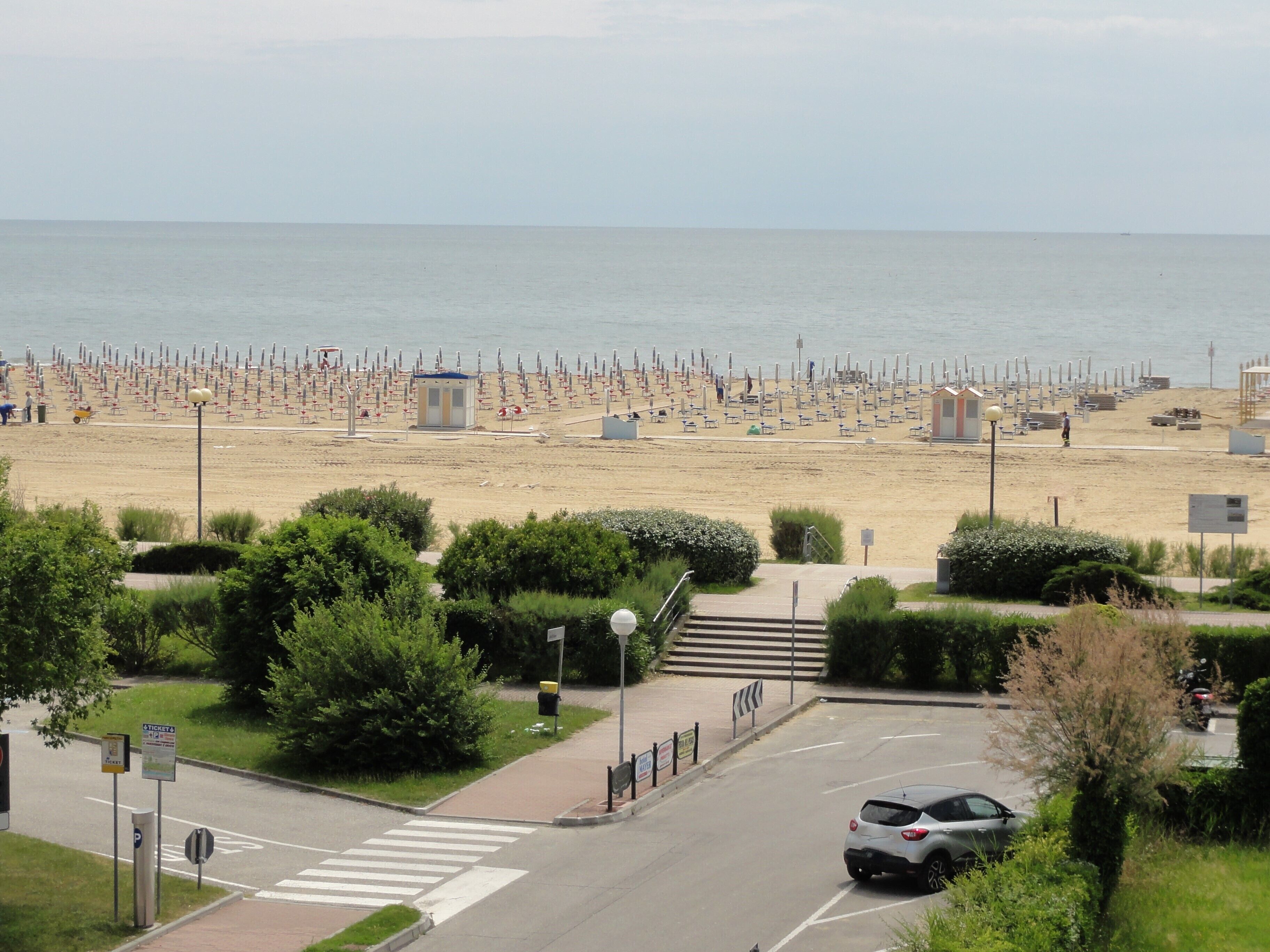 A beach with rows of sunbeds and a small building, a road with a car, and a pedestrian crossing.