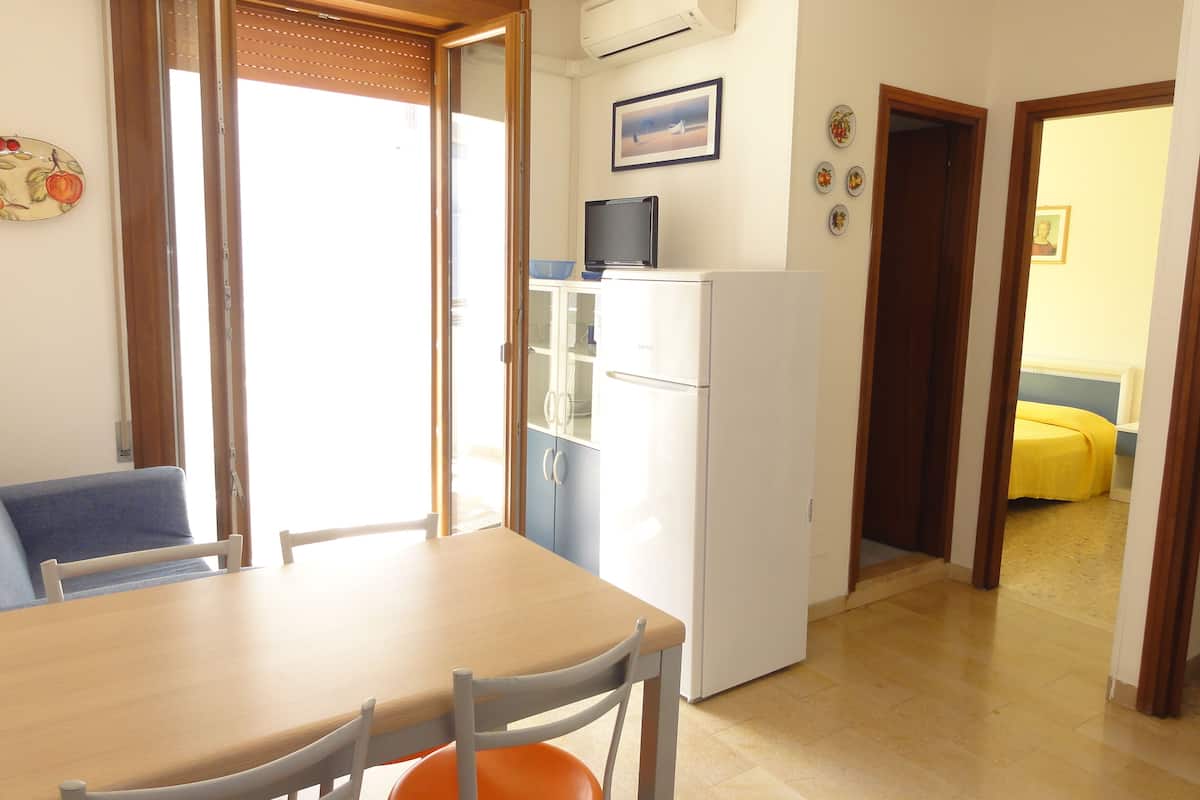 Una cocina compacta con refrigerador, una mesa de comedor con sillas y una puerta corrediza de vidrio que da a un balcón.