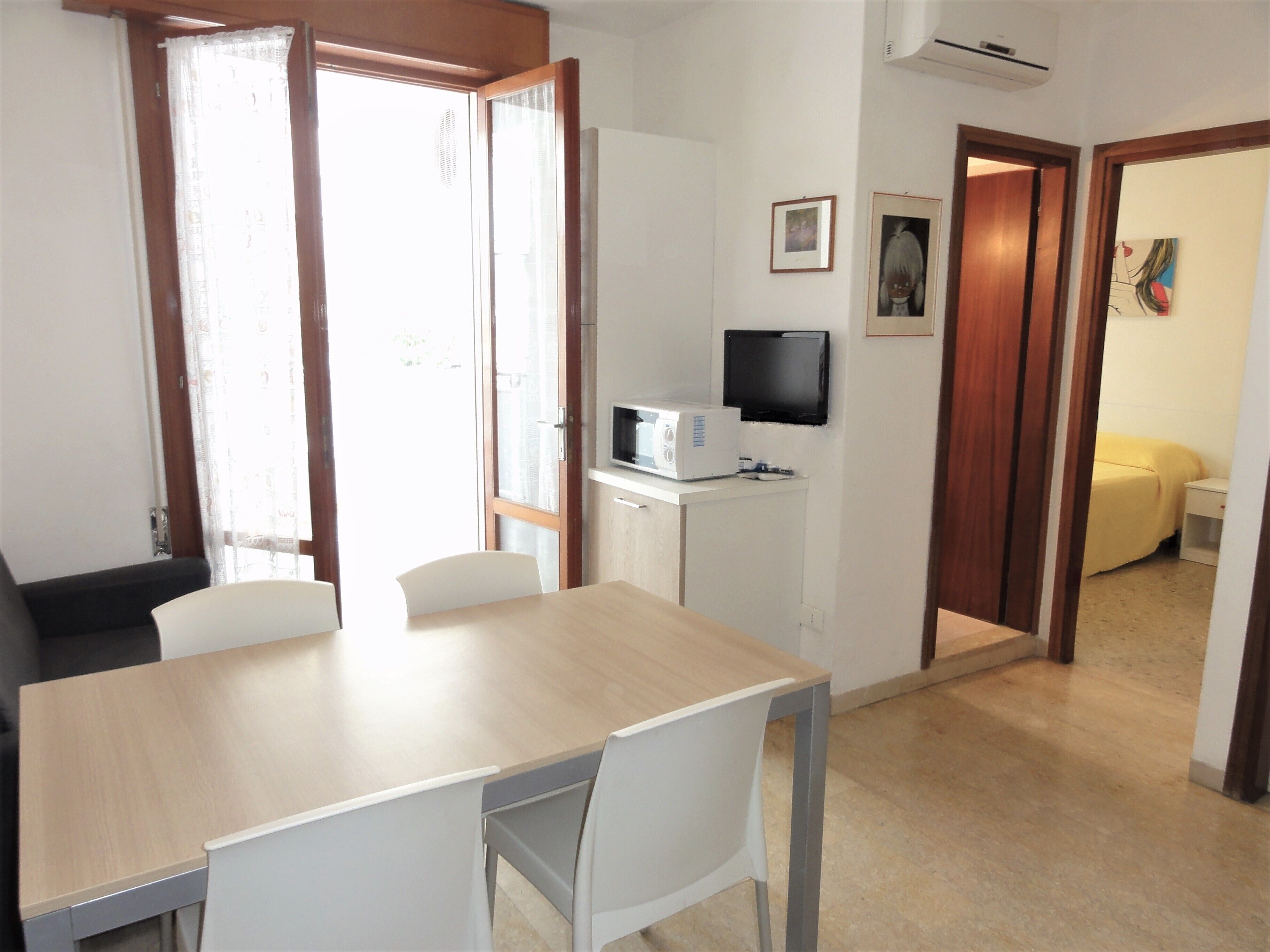 Quarto com mesa de madeira, cadeiras brancas, micro-ondas, televisão e cama com cobertura amarela.