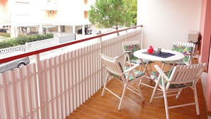 Un balcón con sillas y una mesa blancos, con vista a una calle con coches aparcados y edificios.