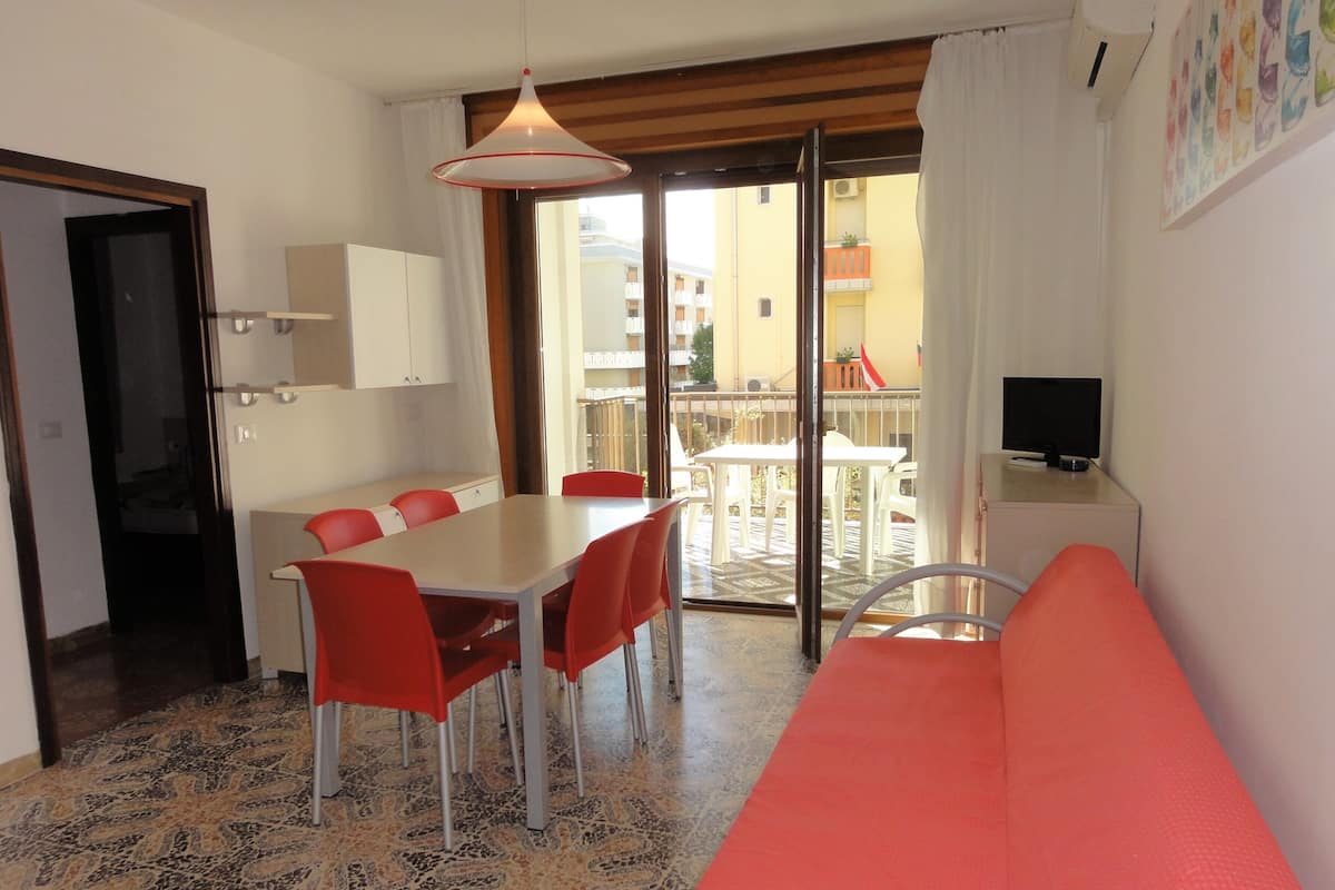 A room with a dining table, red chairs, a sofa, a TV, and a balcony with outdoor furniture.