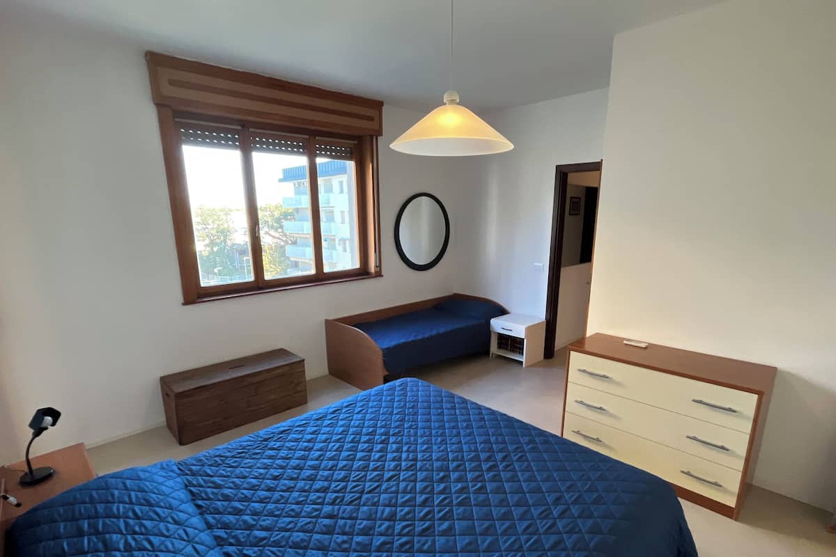 A bedroom with a large bed, a bedside table, a dresser, a window with blinds, and a hanging light fixture.